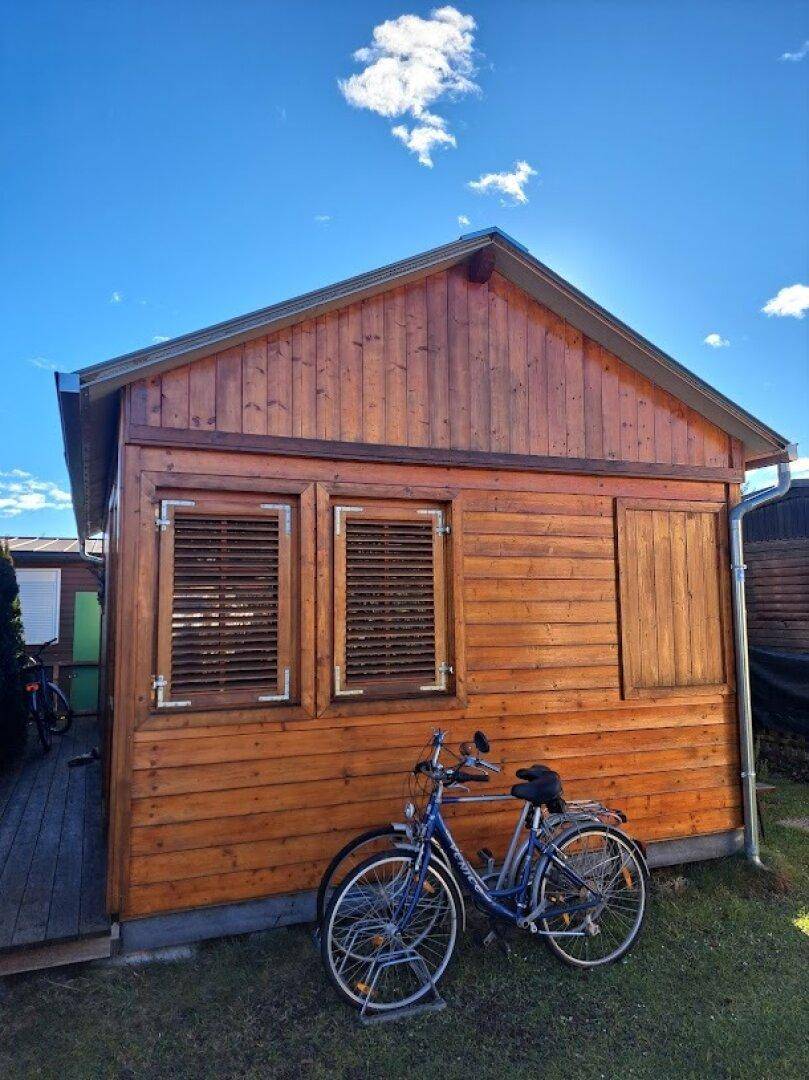 Giebelseite des Holzhauses mit Holzverkleidung, geschlossenen Fensterläden und Fahrrädern im Gras.