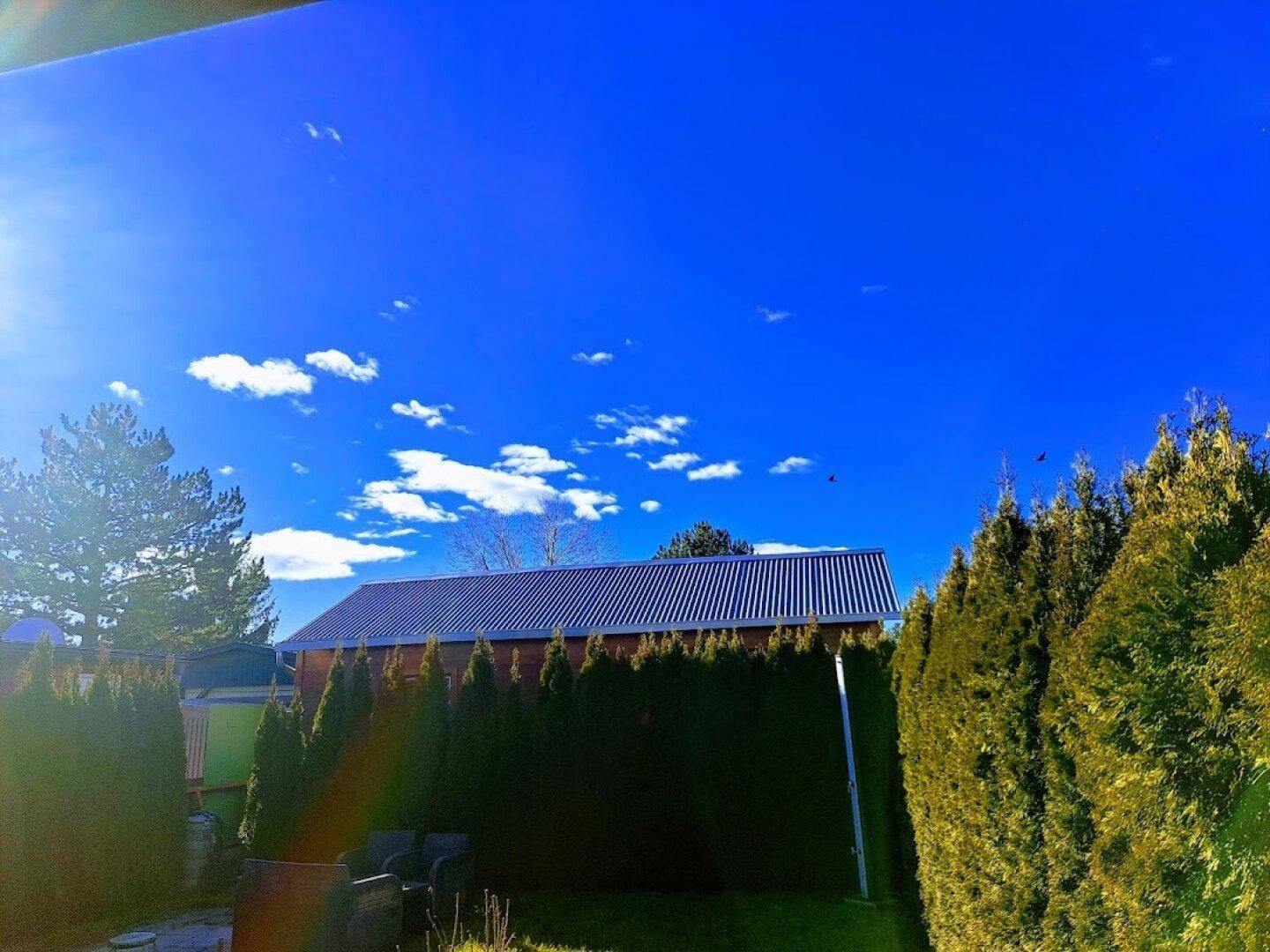 Blick auf ein Gebäude mit Blechdach, umgeben von hohen Hecken unter blauem Himmel.