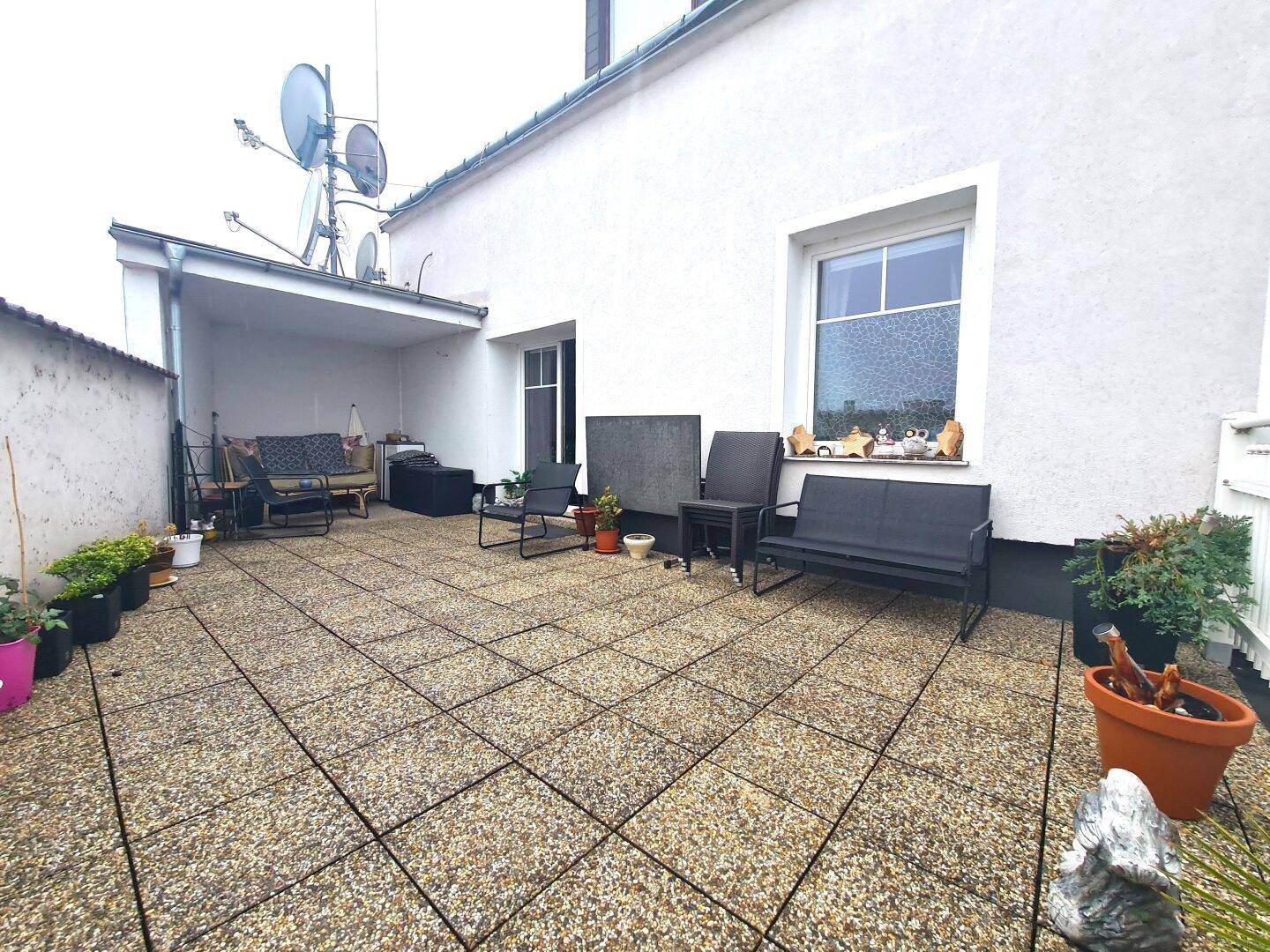 Große Außenterrasse mit Fliesenboden, Sitzgelegenheiten und Topfpflanzen, teilweise überdacht.