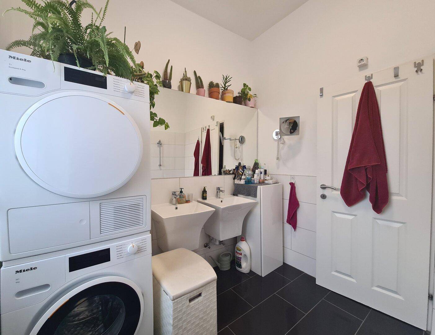 Modernes Badezimmer mit Doppelwaschbecken, Waschmaschine, Trockner und dunklen Bodenfliesen.