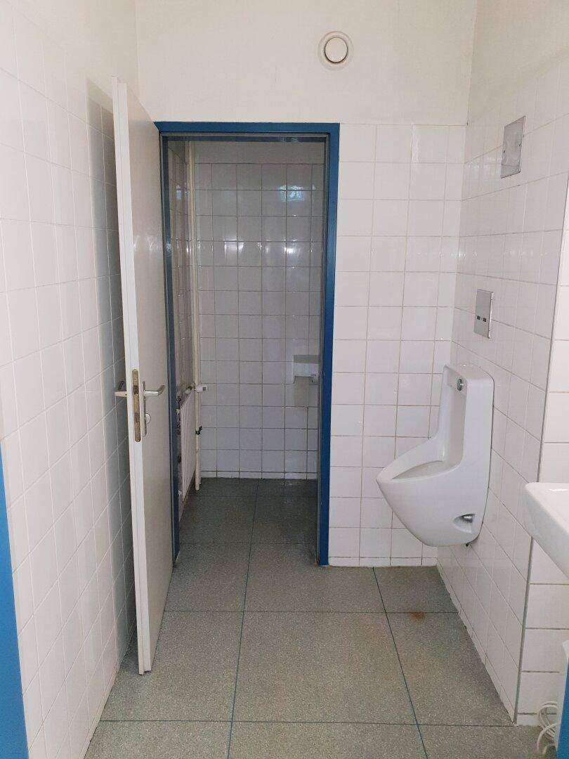 Einfacher Sanitärbereich mit weißen Wandfliesen, einem Urinal und einer Tür zu einem weiteren Raum.