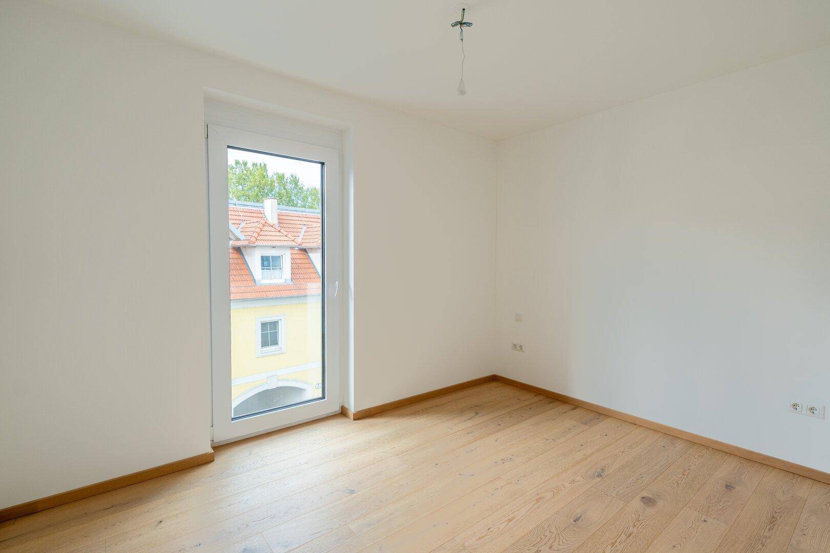 Leeres Zimmer mit hellem Holzboden, weißen Wänden und Fenster mit Blick auf ein Gebäude.