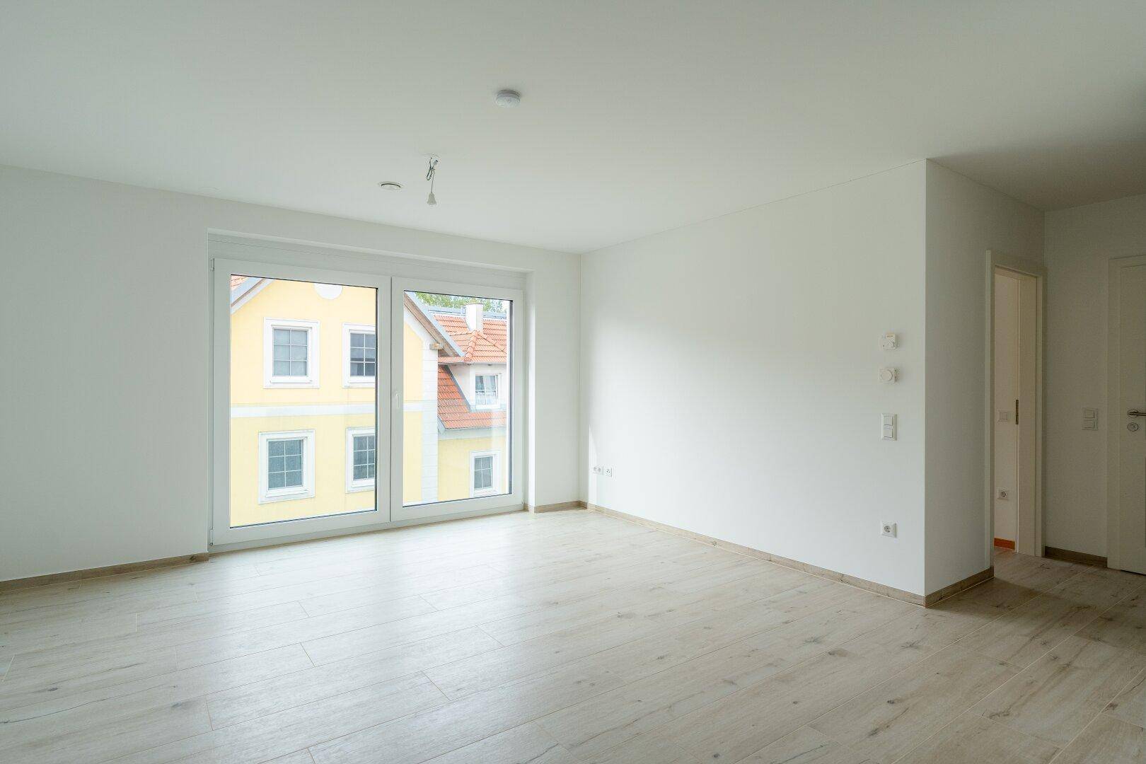 Helles, leeres Zimmer mit großen Fenstern, hellem Holzboden und Blick auf ein Nachbargebäude.