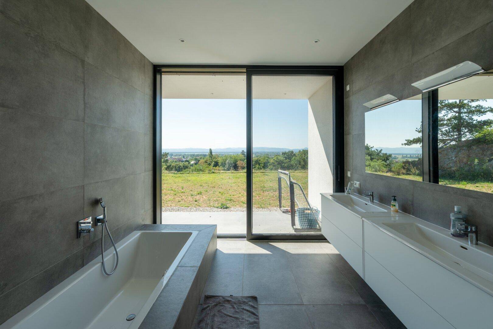 Modernes Badezimmer mit Badewanne, Doppelwaschbecken und großer Schiebetür mit Landschaftsblick.
