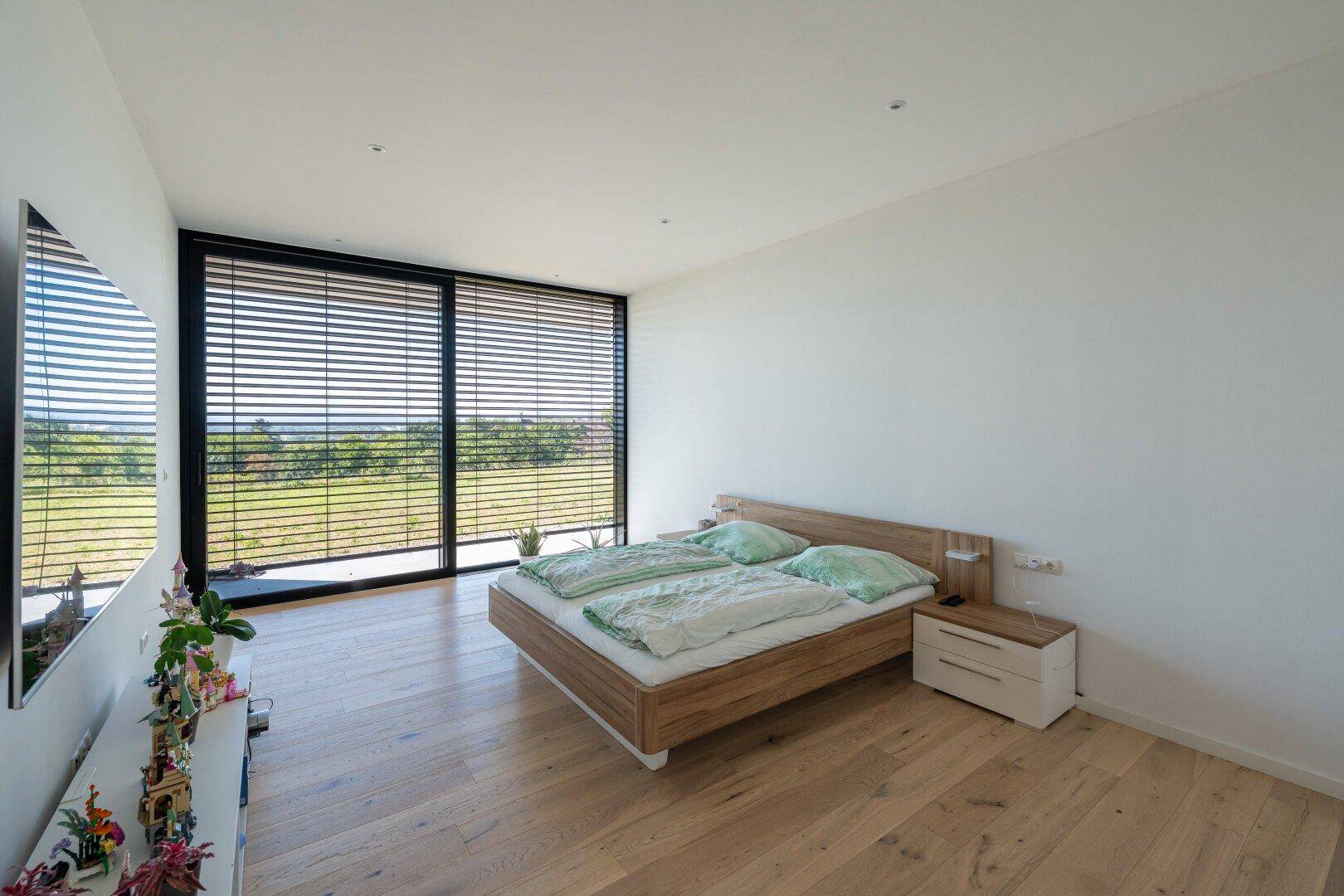 Schlafzimmer mit Holzbett, Nachttisch und großem Fenster mit Jalousien und grüner Aussicht.