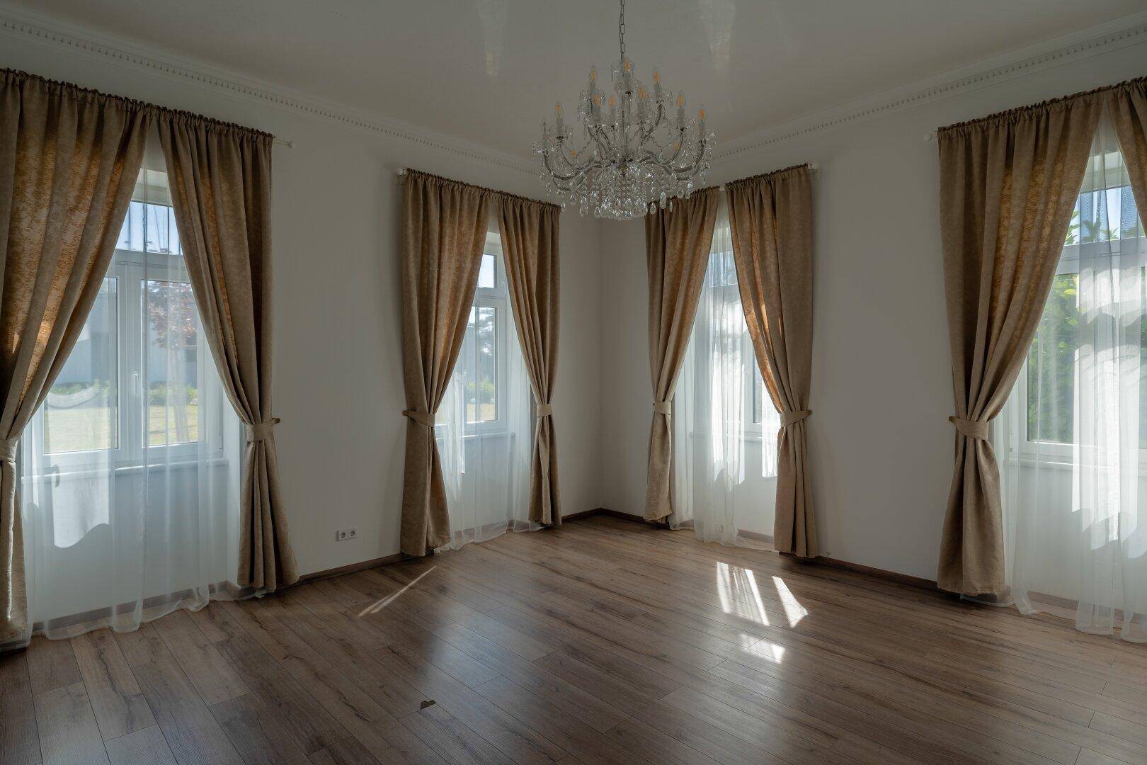 Heller Raum mit mehreren Fenstern, beigen Vorhängen, Kronleuchter und Holzboden.