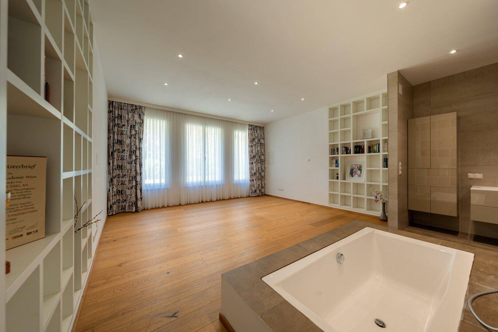 Geräumiger Raum mit Holzboden, Einbauregalen und integrierter Badewanne, hell durch Fenster.