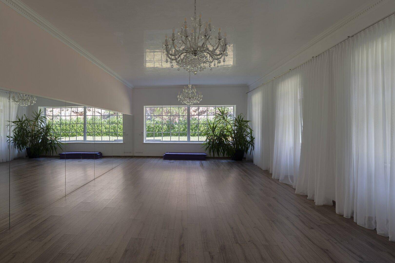 Großer Raum mit Spiegelwänden, vielen Fenstern, Kronleuchtern und Holzboden, ideal für Sport.