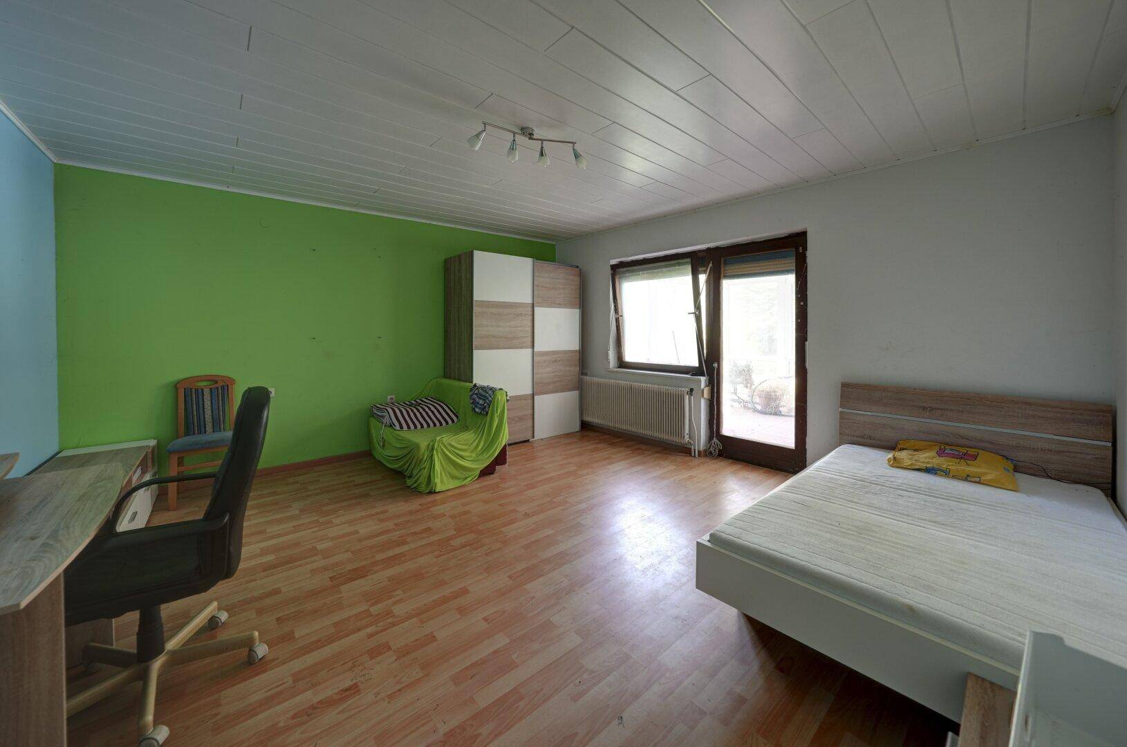 Schlafzimmer mit Laminatboden, Bett, Schreibtisch und einem Kleiderschrank, eine Wand ist grün gestrichen.