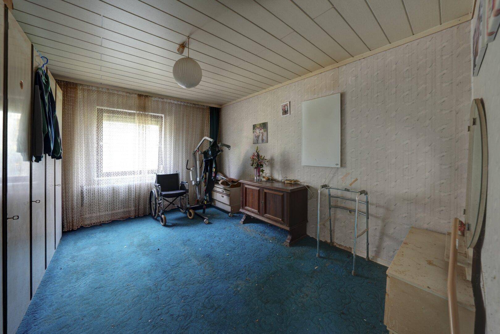Schlafzimmer mit blauem Teppichboden und Hilfsmitteln, das auf Barrierefreiheit hindeutet.