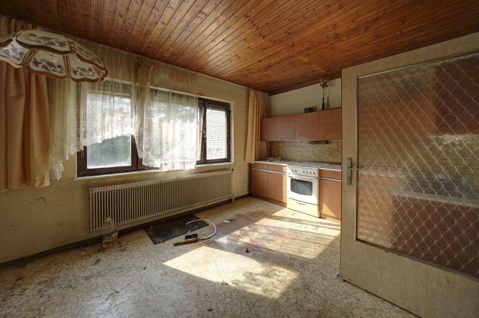 Küche mit Fenster, Heizkörper und älteren Holzschränken, die eine Renovierung benötigt.