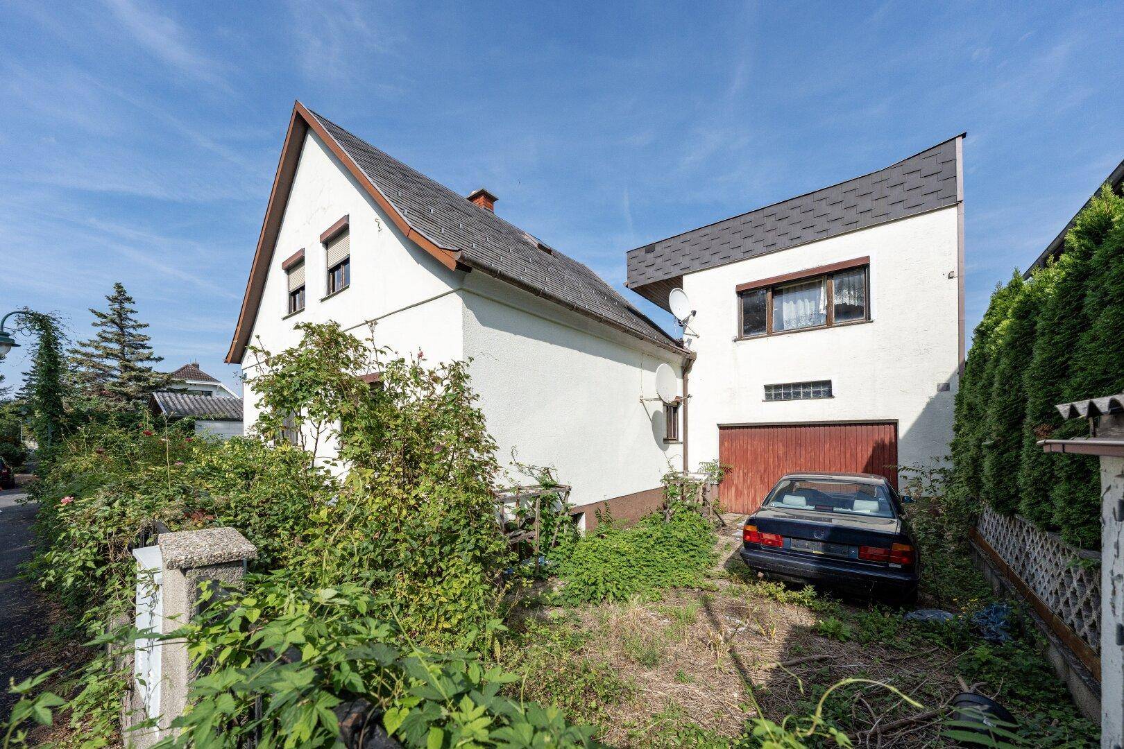 Ansicht eines Einfamilienhauses mit Satteldach, einer Einfahrt und üppiger Vegetation im Vorgarten.