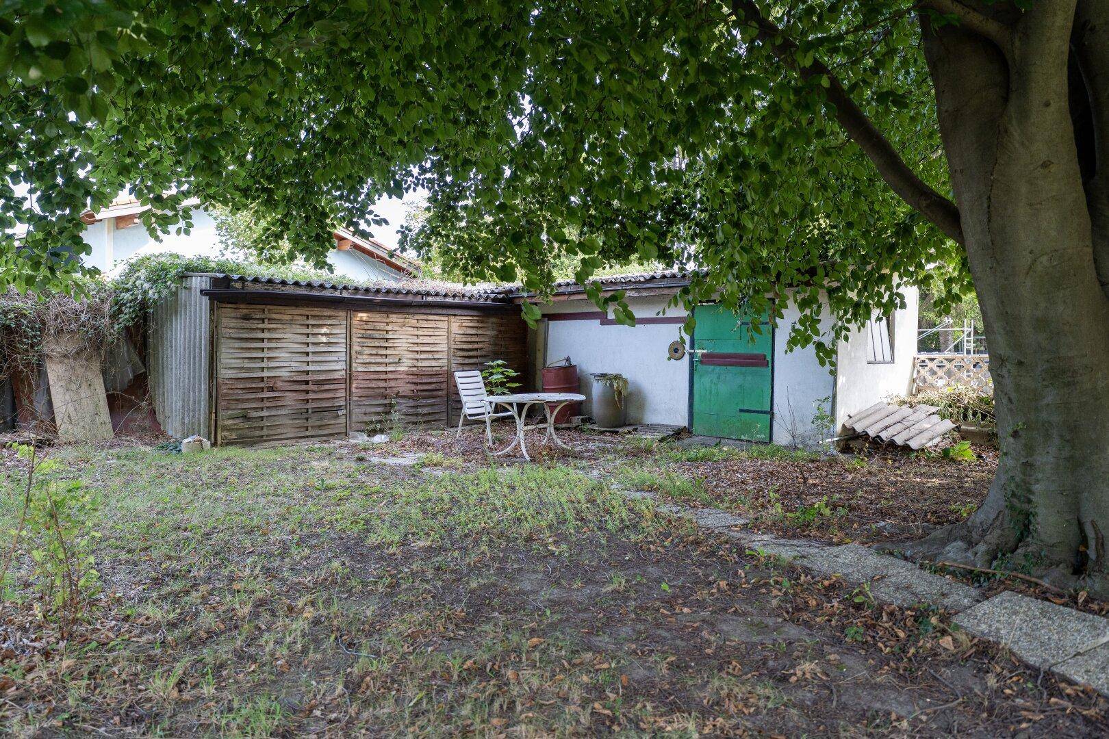 Gartenbereich mit einem Schuppen, Holzzaun und einer kleinen Sitzgelegenheit unter Bäumen.