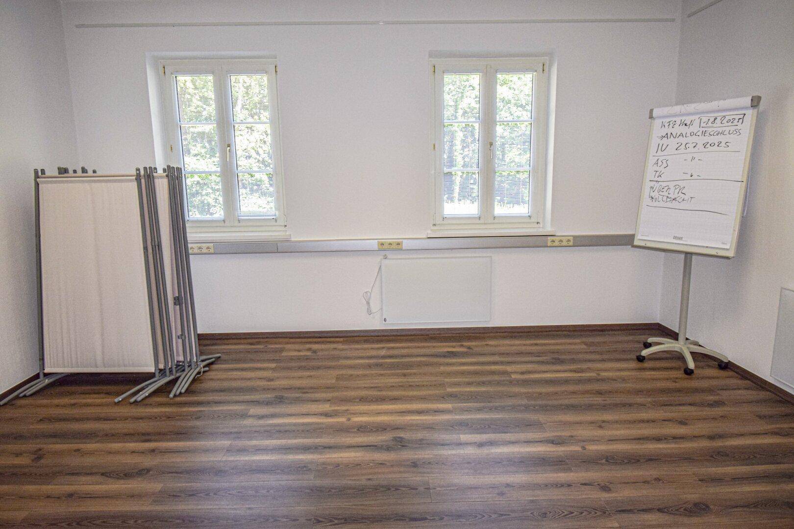 Leerer Raum mit Laminatboden, zwei Fenstern und einer Flipchart, ideal für Präsentationen oder Schulungen.