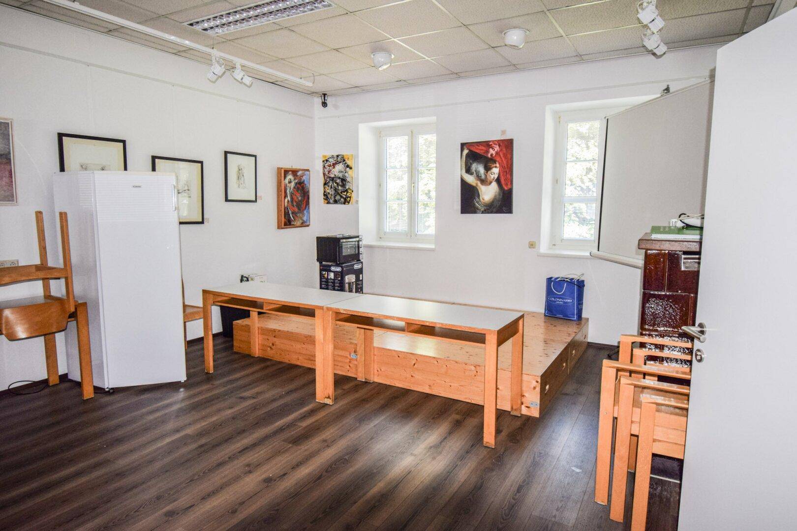 Heller Mehrzweckraum mit Laminatboden, einem Kühlschrank und einer erhöhten Holzplattform für Ausstellungen.