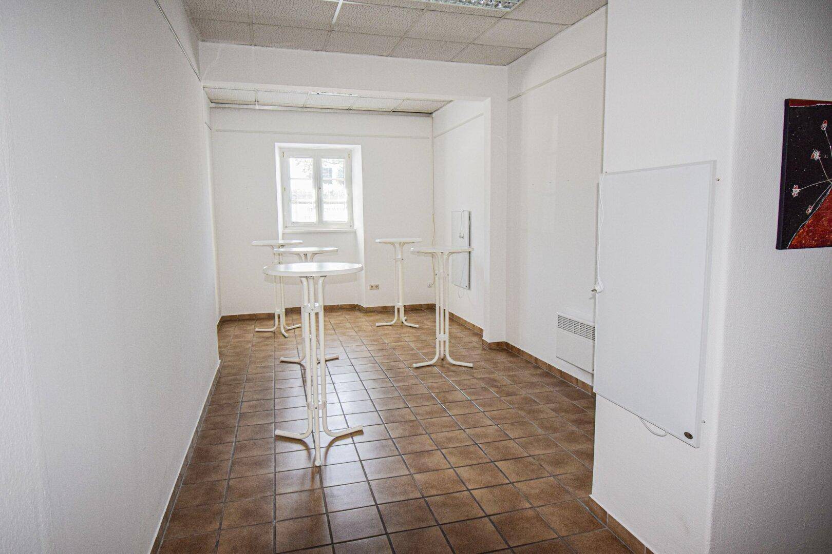 Kleinerer Raum mit braunen Fliesen, Stehtischen und einem Fenster, geeignet für Empfänge oder Pausenbereiche.