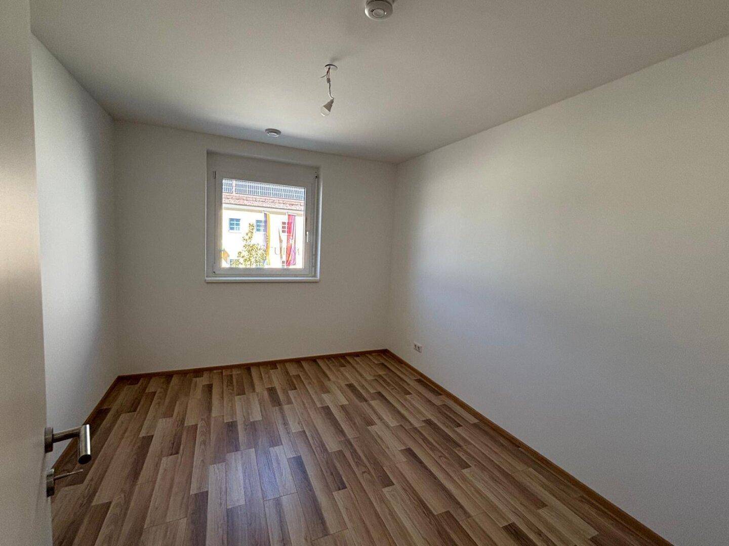 Helles, unmöbliertes Zimmer mit Laminatboden und einem Fenster mit Blick nach draußen.