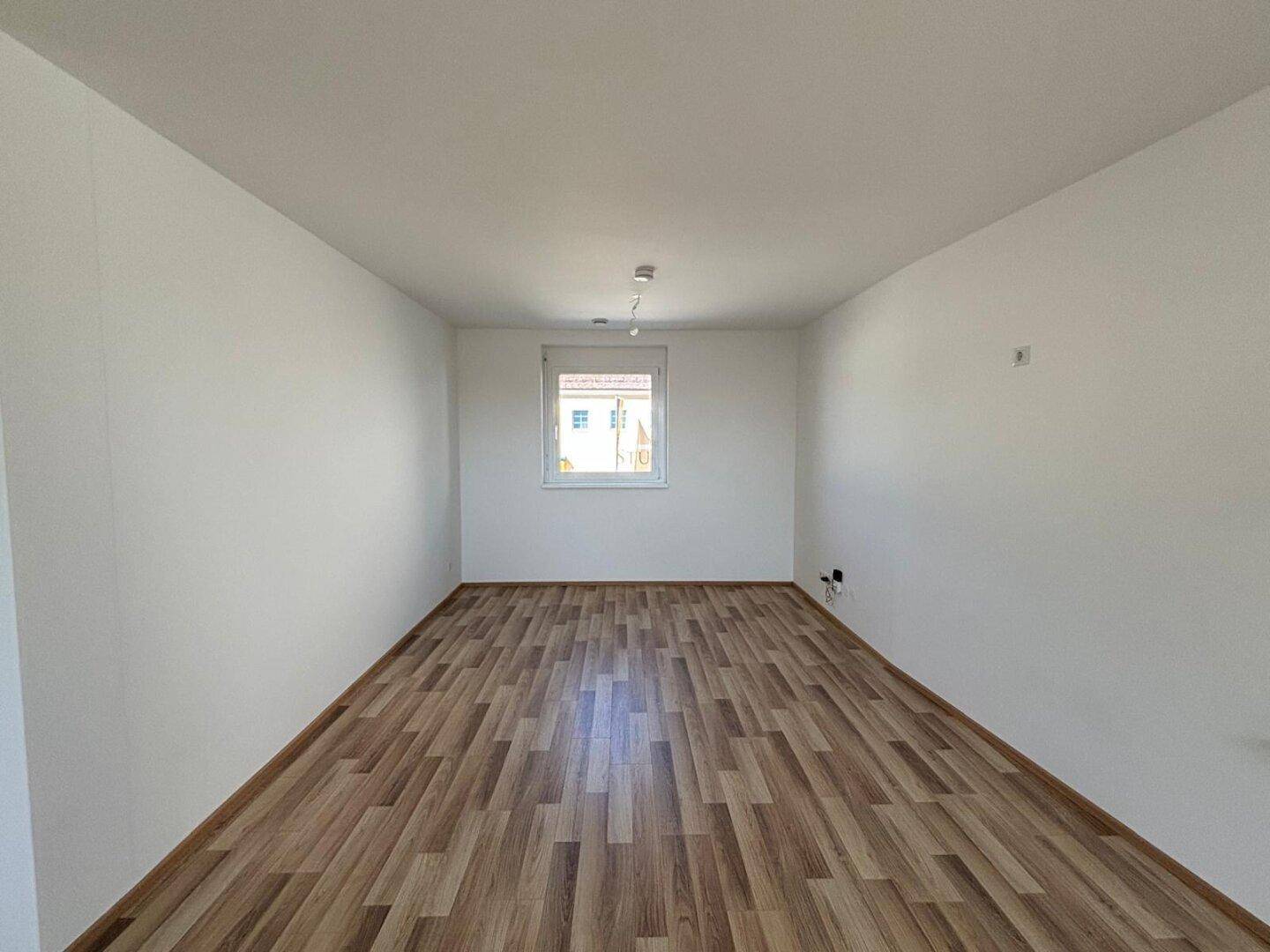Geräumiges, leeres Zimmer mit Laminatboden und einem einzelnen Fenster für Tageslicht.