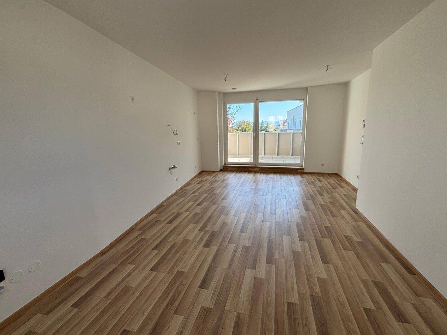 Heller Wohnbereich mit Laminatboden und großen Fenstern, die Zugang zum Balkon bieten.