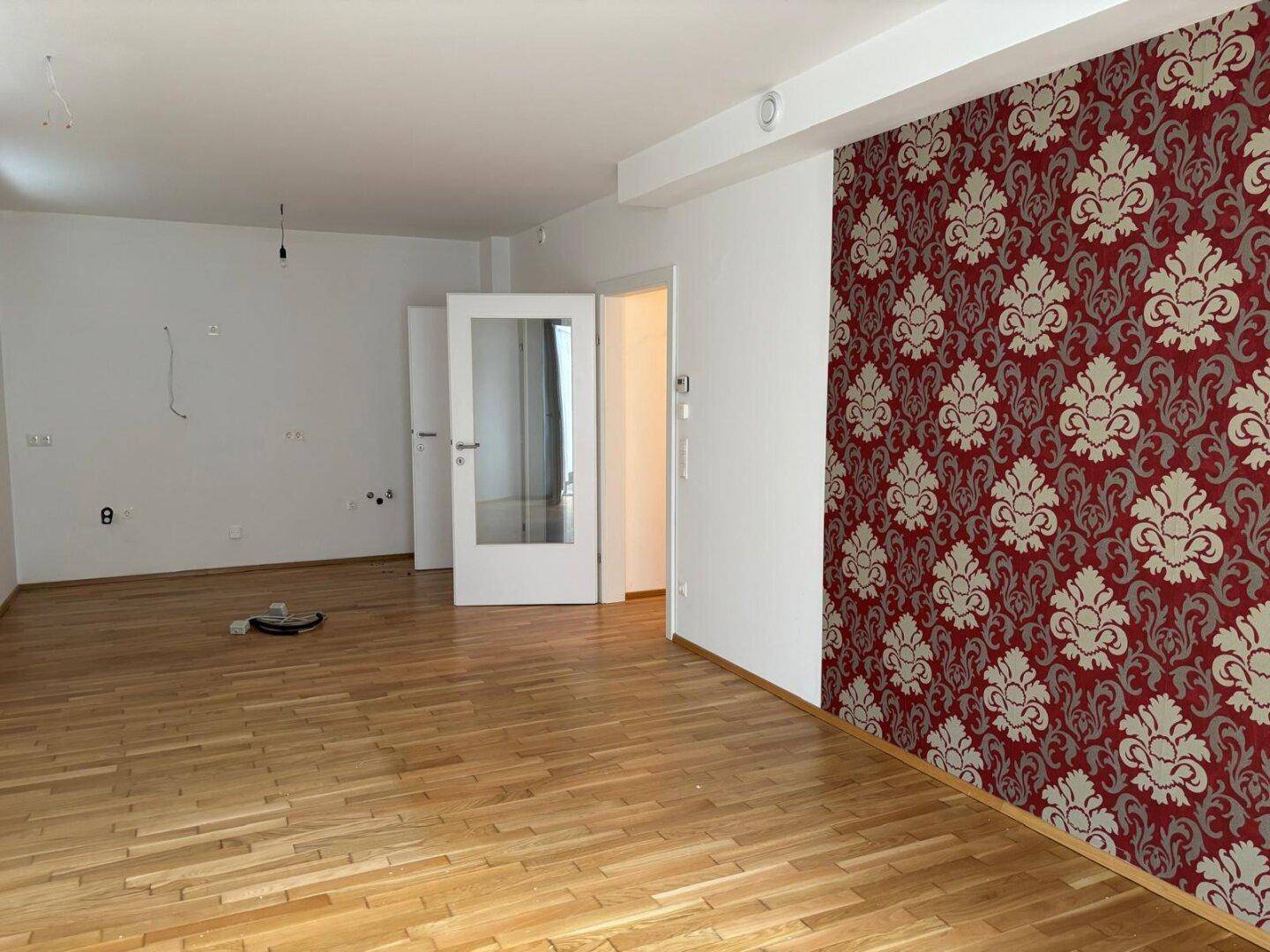 Geräumiger, unmöblierter Wohnbereich mit Holzboden und auffälliger gemusterter Tapetenwand.