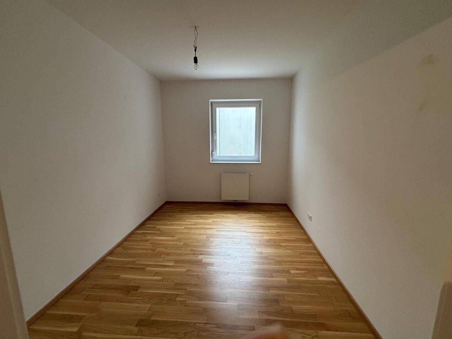 Helles, unmöbliertes Zimmer mit Holzboden, Fenster und Heizkörper an der Wand.