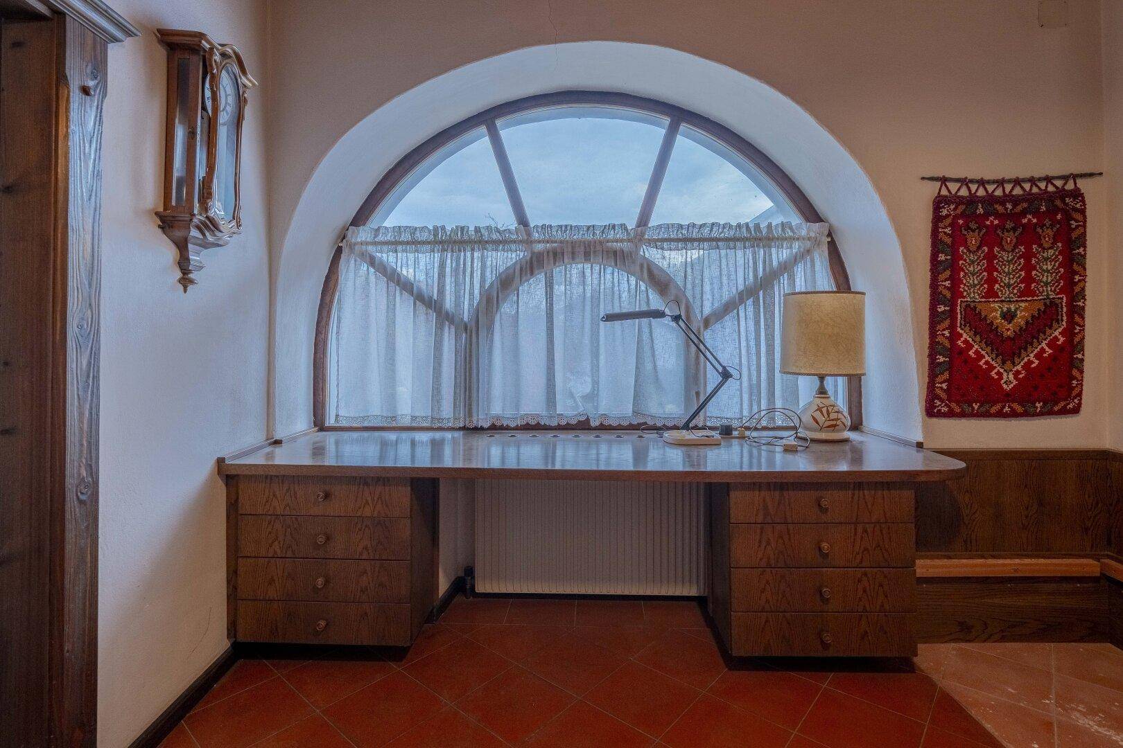 Arbeitsbereich mit großem Schreibtisch unter einem Rundbogenfenster und Terrakottafliesen.