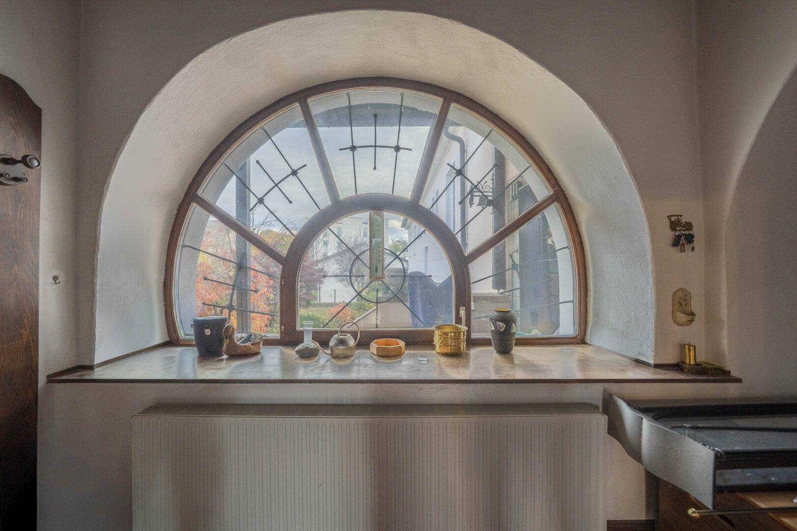 Blick durch ein großes Rundbogenfenster mit Sprossen auf die umliegende Landschaft und den Garten.