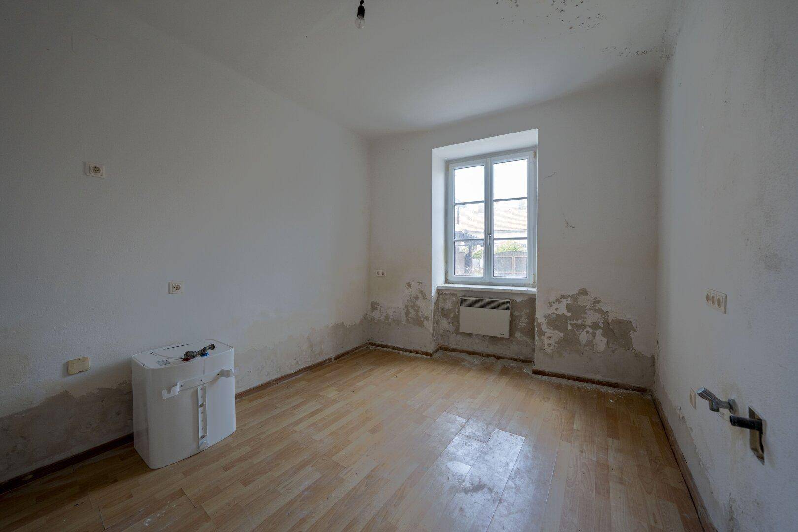 Leeres Zimmer mit abblätternder Farbe, Holzboden und einem Fenster, das Renovierungsbedarf zeigt.