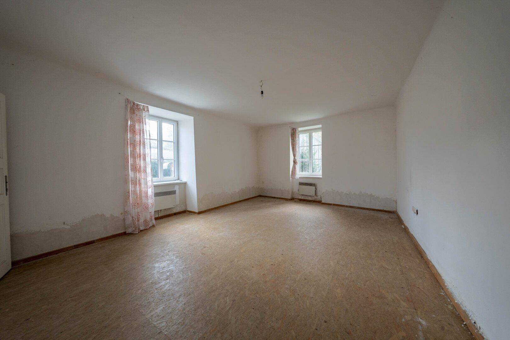 Leeres Zimmer mit zwei Fenstern, einfachen Vorhängen und deutlichem Renovierungsbedarf an den Wänden.