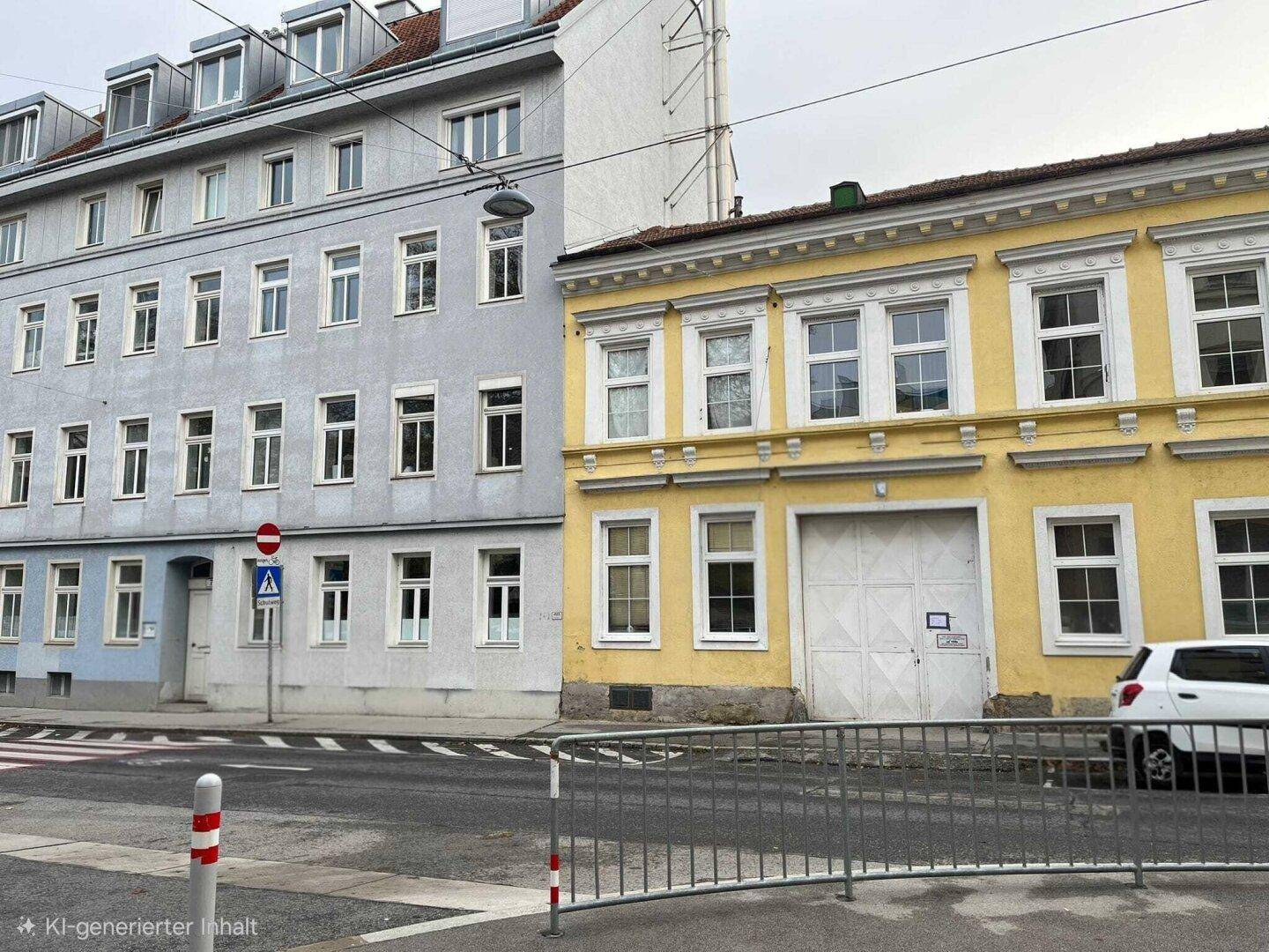 Ansicht der gelben Gebäudefassade an einer Straße, daneben ein graues Nachbargebäude mit vielen Fenstern.