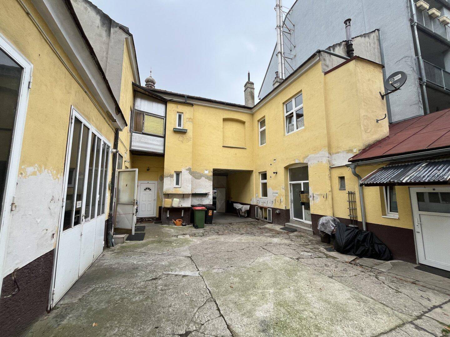Weitläufiger Innenhof mit Betonboden und der Rückseite des gelben Gebäudes, das Renovierung benötigt.