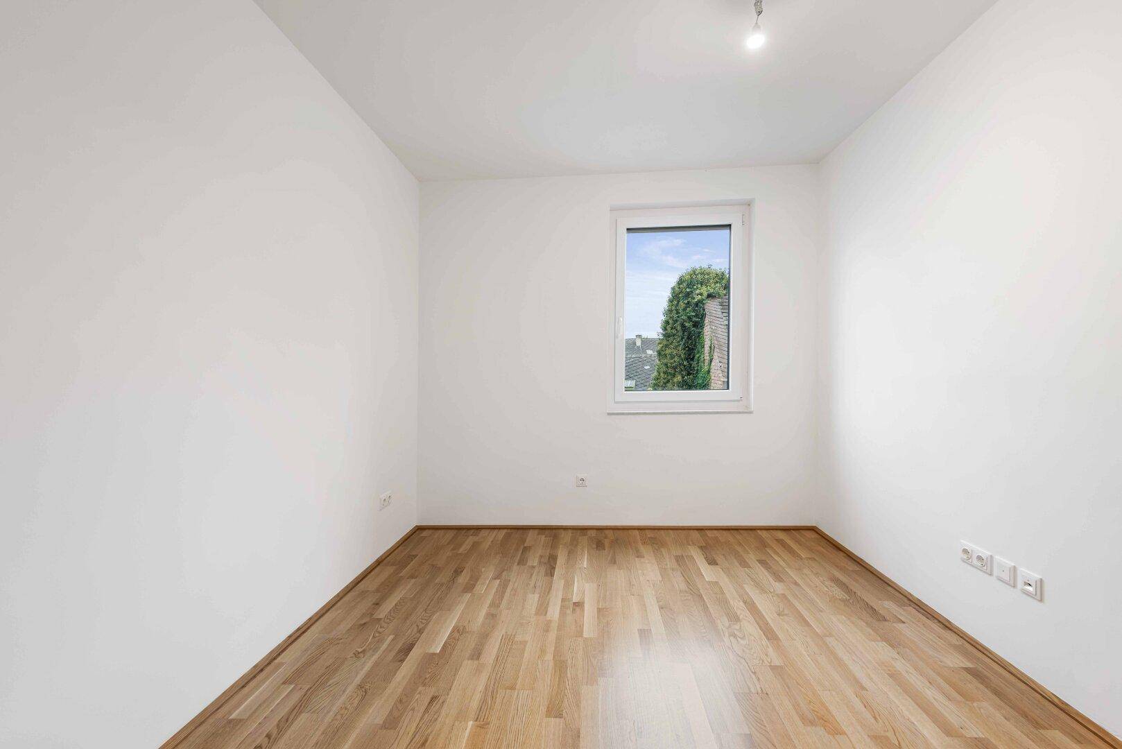 Helles Zimmer mit Parkettboden und einem Fenster mit Blick ins Grüne.