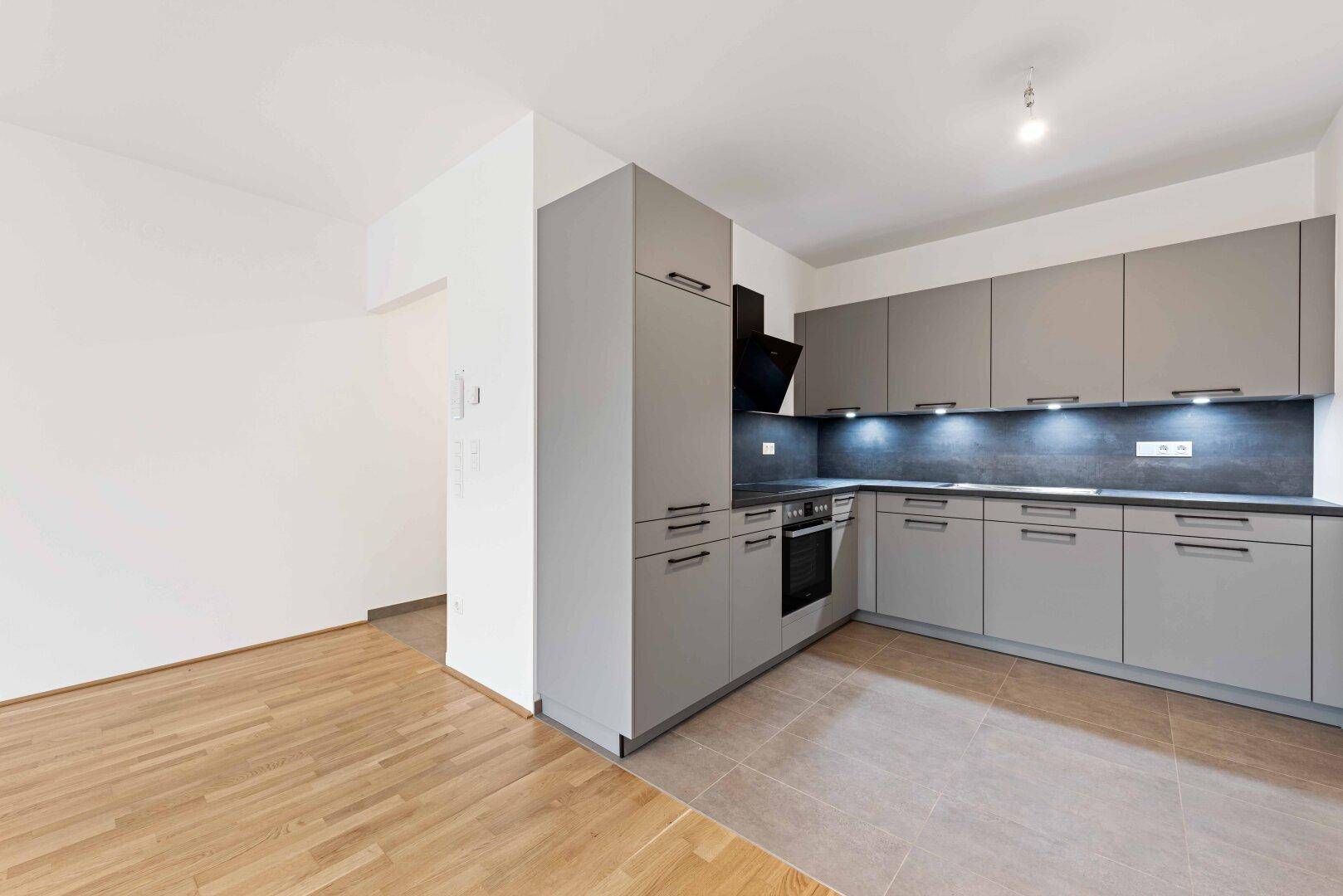 Helle Küche mit grauen Schränken und angrenzendem Raum mit Holzparkettboden.