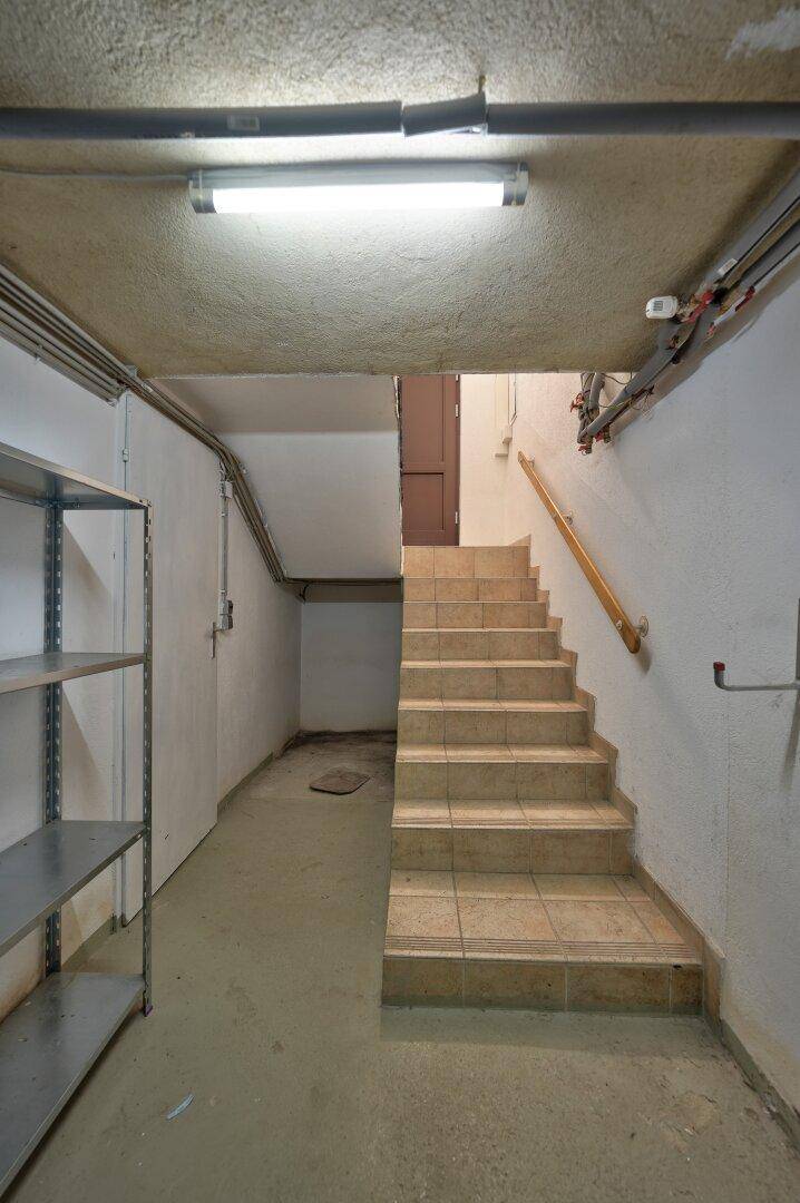 Kellerflur mit Treppenaufgang, Regalen und sichtbaren Versorgungsleitungen an der Decke.