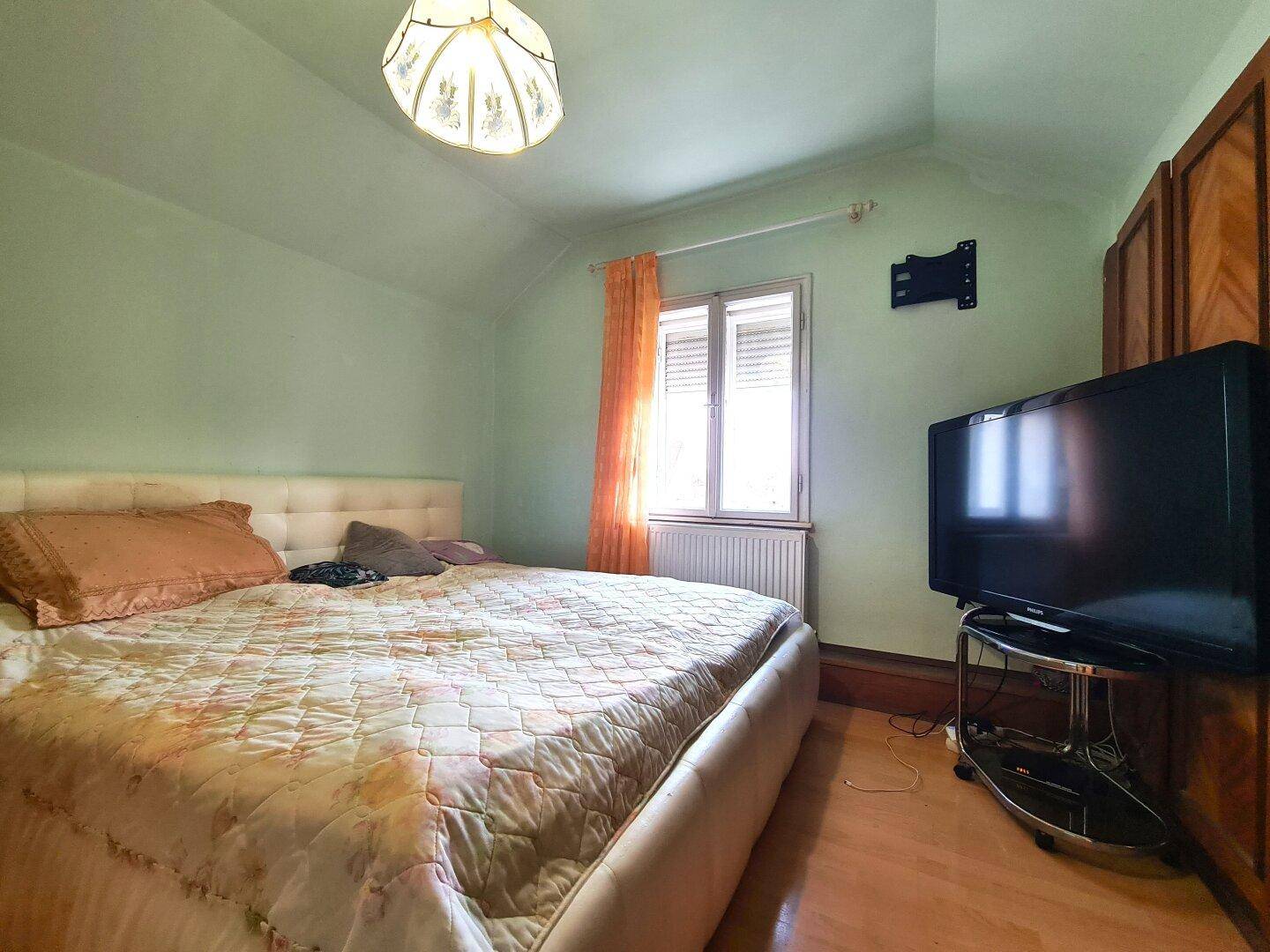 Schlafzimmer mit Doppelbett, Holzschrank, Fernseher und Fenster mit Jalousien.