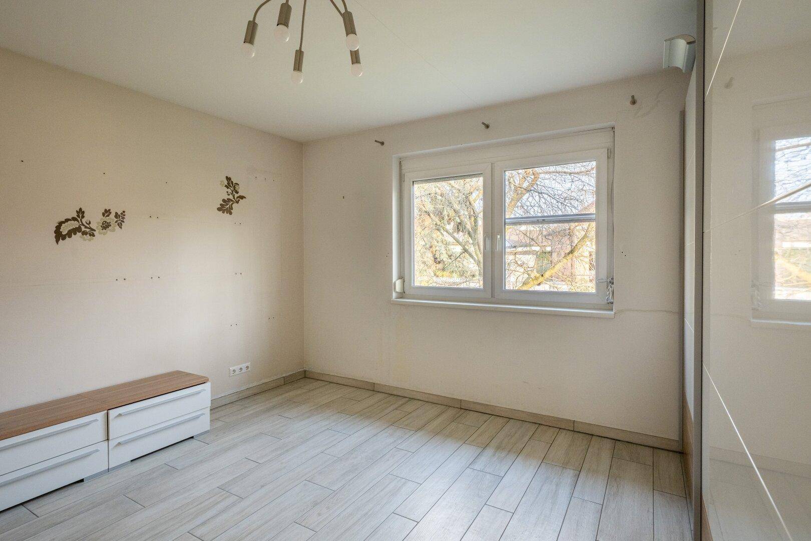 Helles Schlafzimmer mit Fliesenboden in Holzoptik und einem Fenster mit Blick ins Grüne.