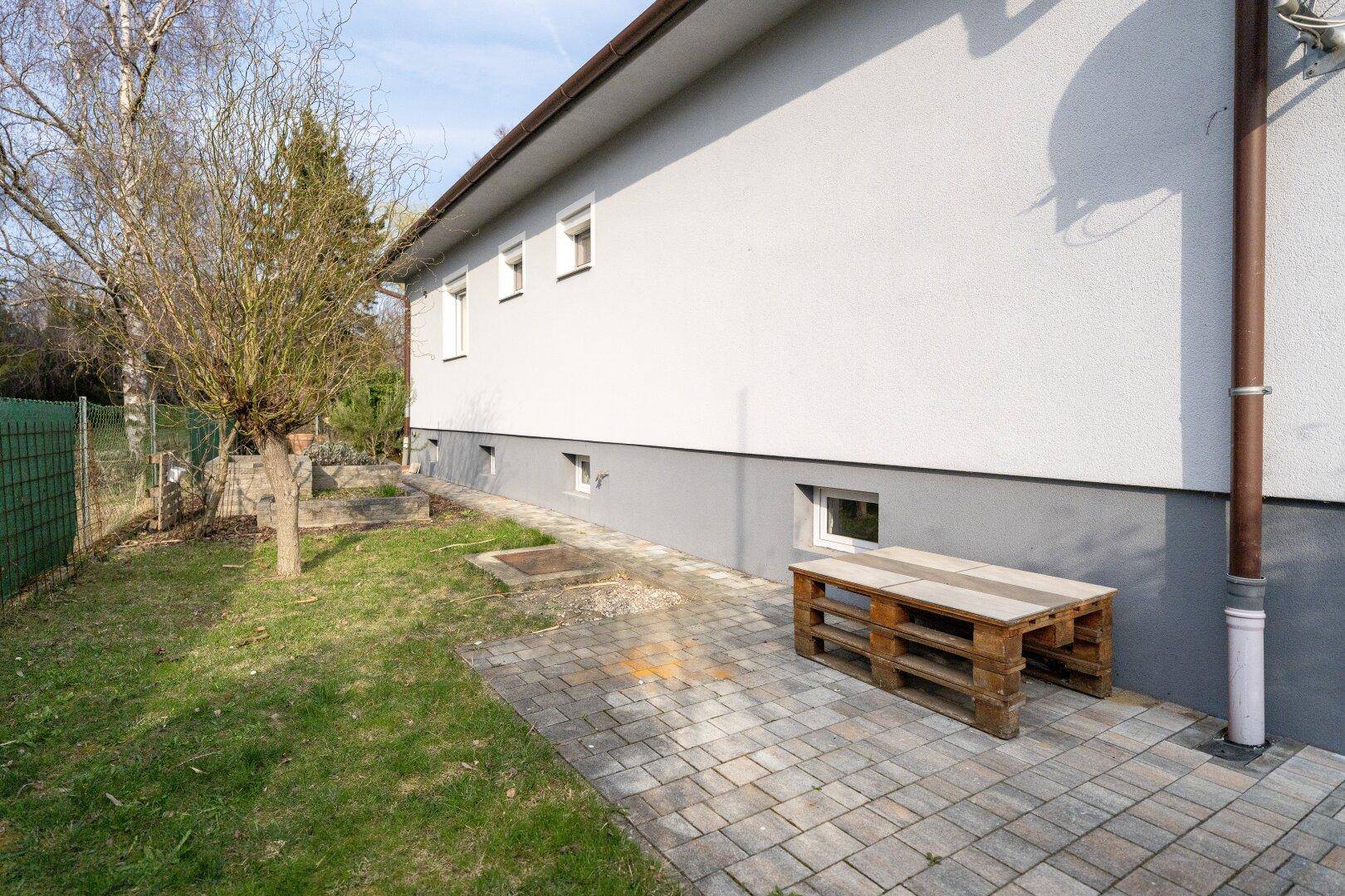 Seitenansicht des Hauses mit kleinem gepflasterten Bereich und einer einfachen Holzbank im Garten.