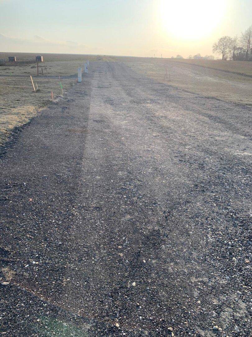 Langer Schotterweg durch ein weites Feld unter einem hellen Himmel bei Sonnenuntergang.