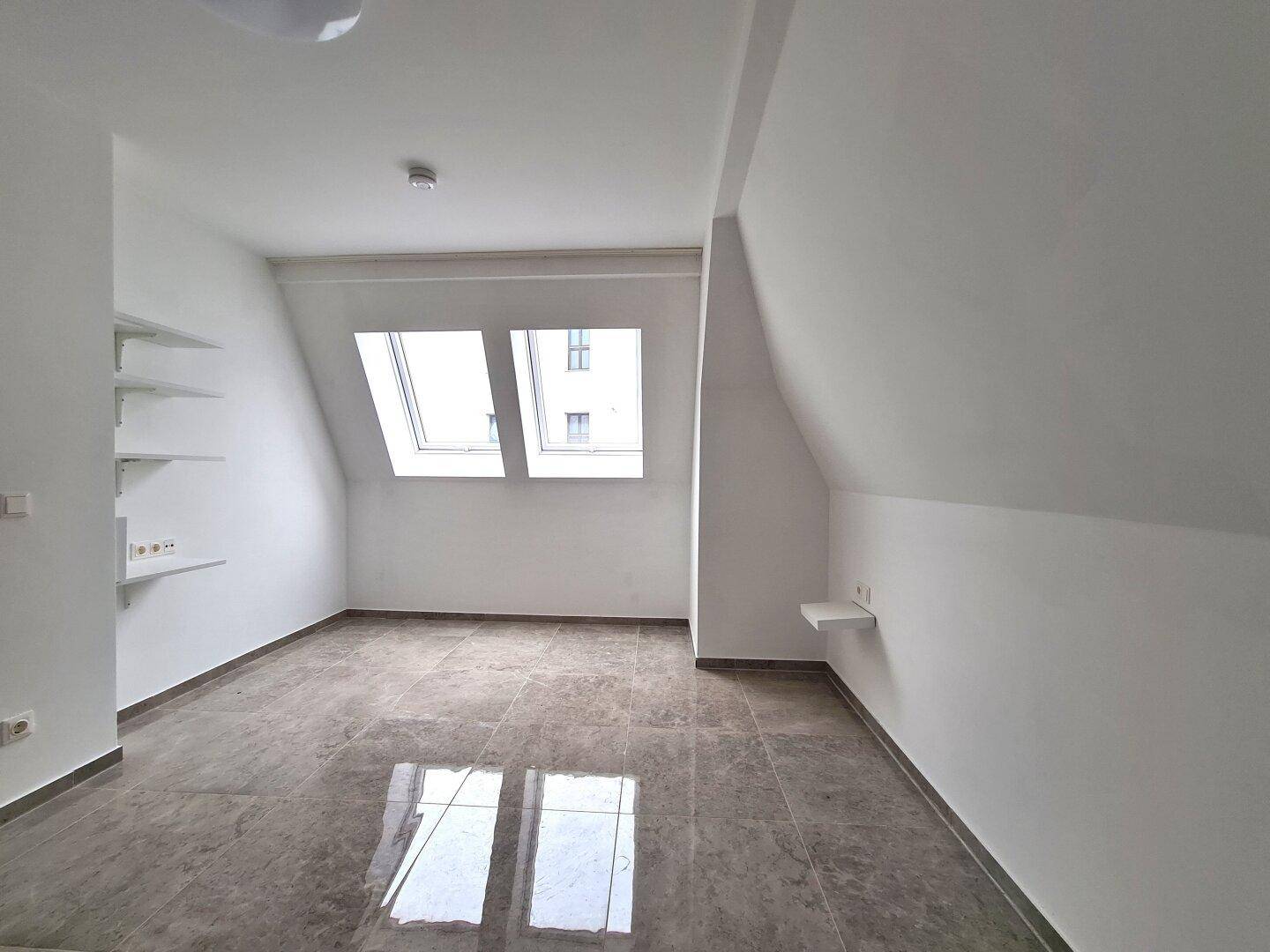 Heller Raum im Dachgeschoss mit schrägen Wänden, Dachfenstern und glänzendem Fliesenboden.