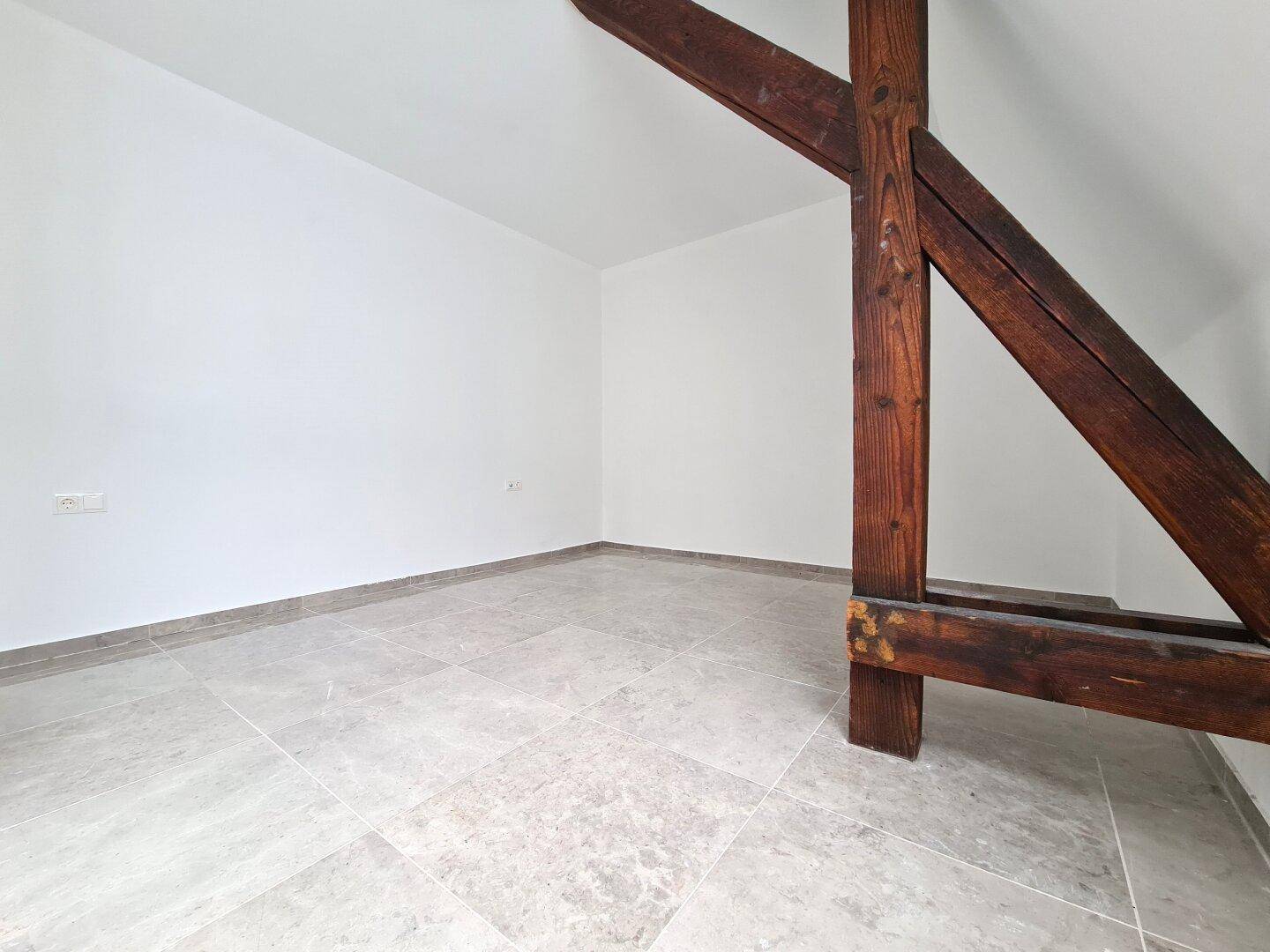 Leerer Raum mit schrägen Wänden, Holzbalken und hellem Fliesenboden.