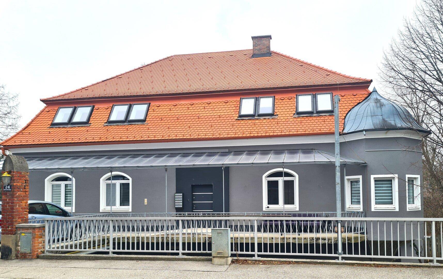 Graues Gebäude mit orangefarbenem Ziegeldach, Gaubenfenstern und einer Rampe am Eingang.