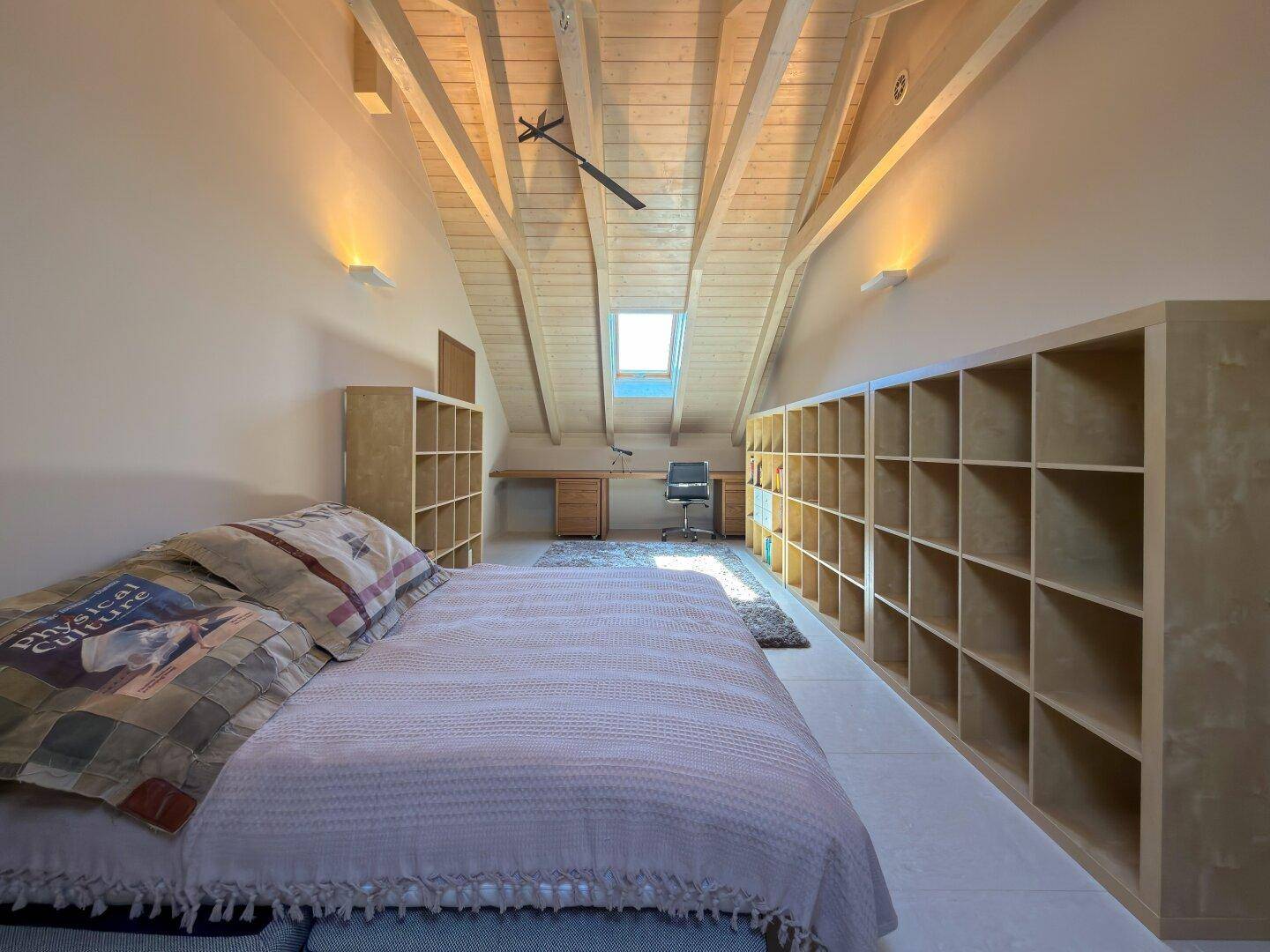 Helles Schlafzimmer im Dachgeschoss mit Regalen, Schreibtisch und Dachfenster.