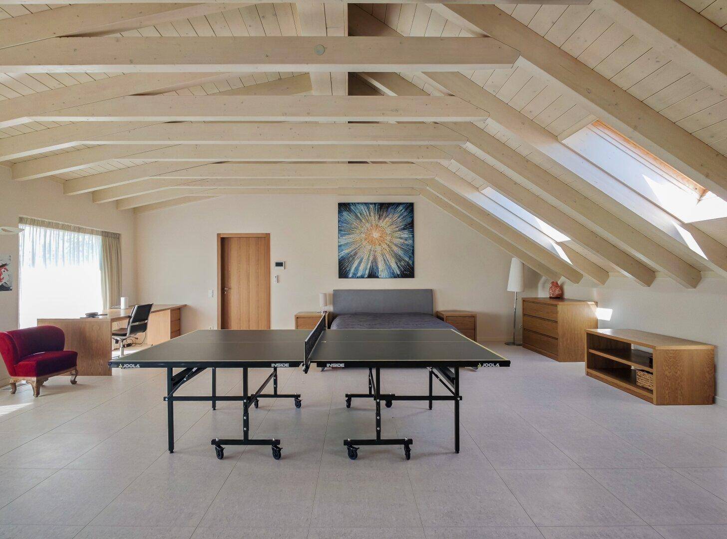 Geräumiger Dachboden mit Tischtennisplatte, Schlafbereich und Schreibtisch unter Holzbalken.