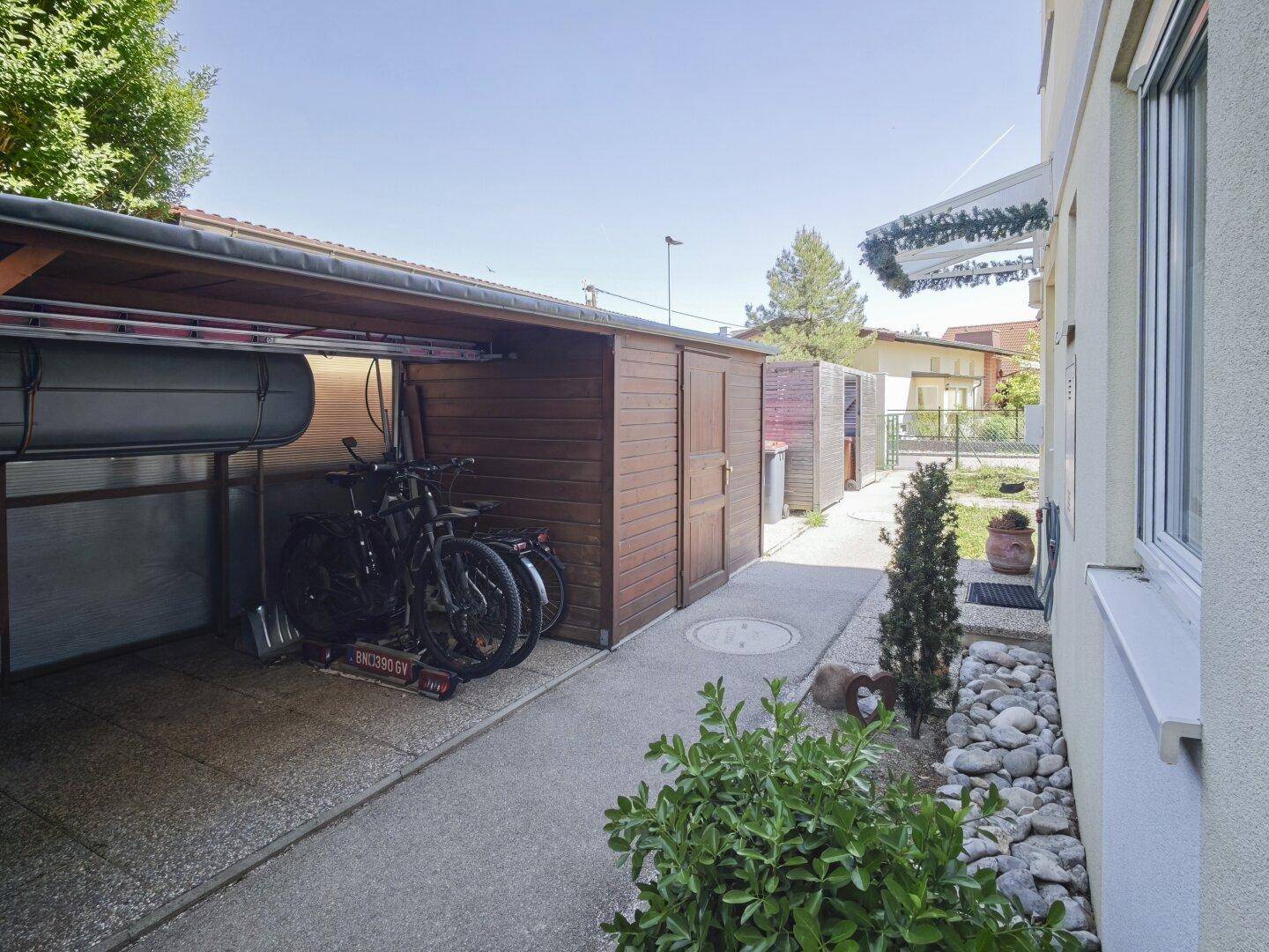 Carport mit Fahrrädern und angrenzendem Schuppen, umgeben von Grünflächen.