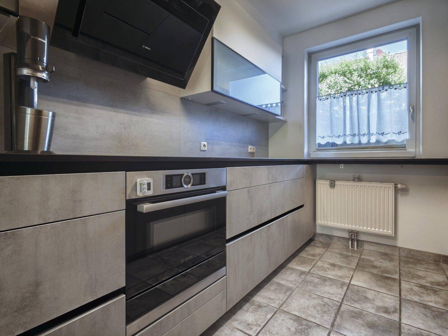 Moderne Küche mit grauen Fronten, integriertem Backofen und Fenster für Tageslicht.