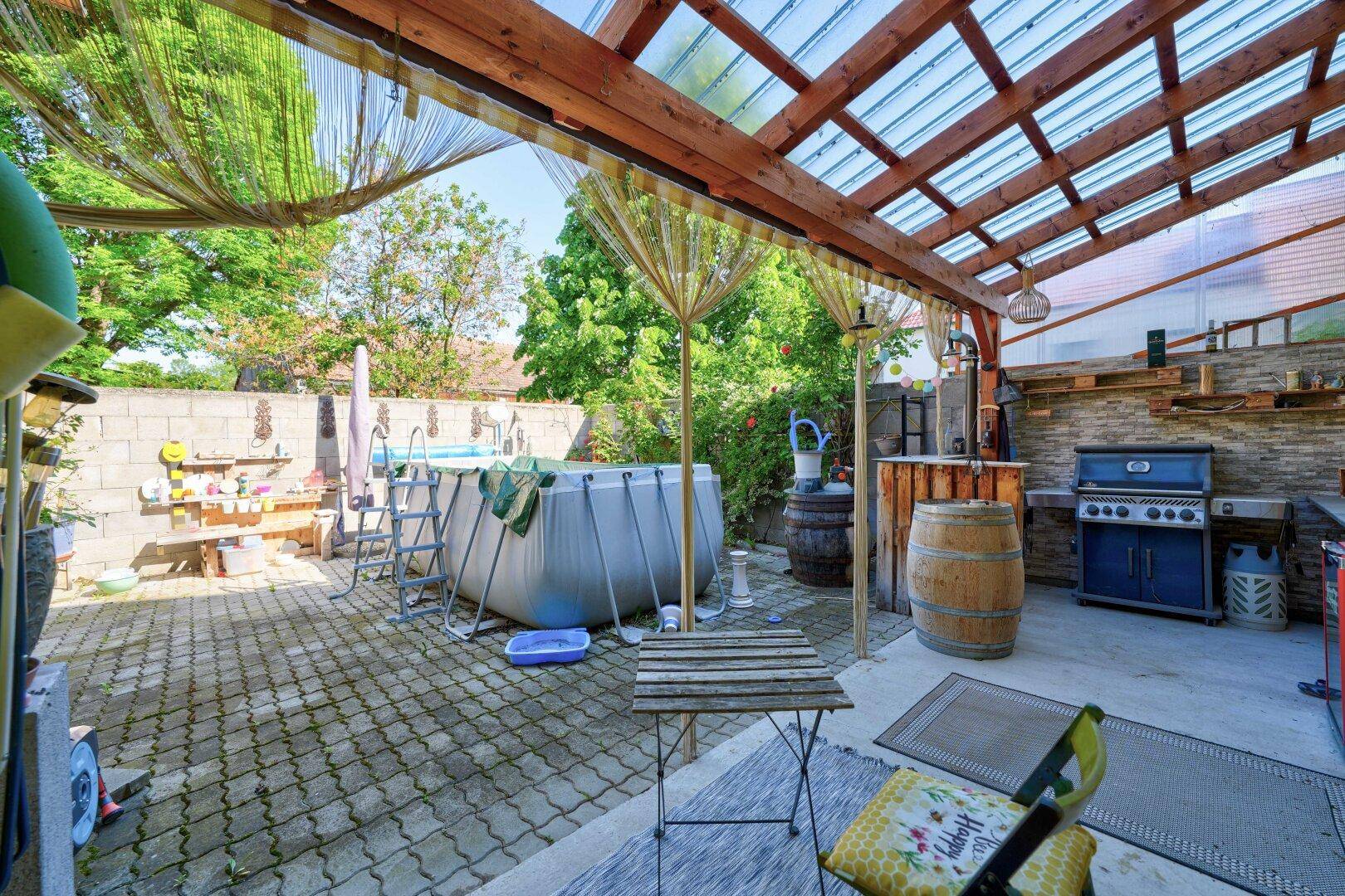 Überdachte Terrasse mit Grillbereich, Sitzgelegenheiten und einem Aufstellpool im Garten.