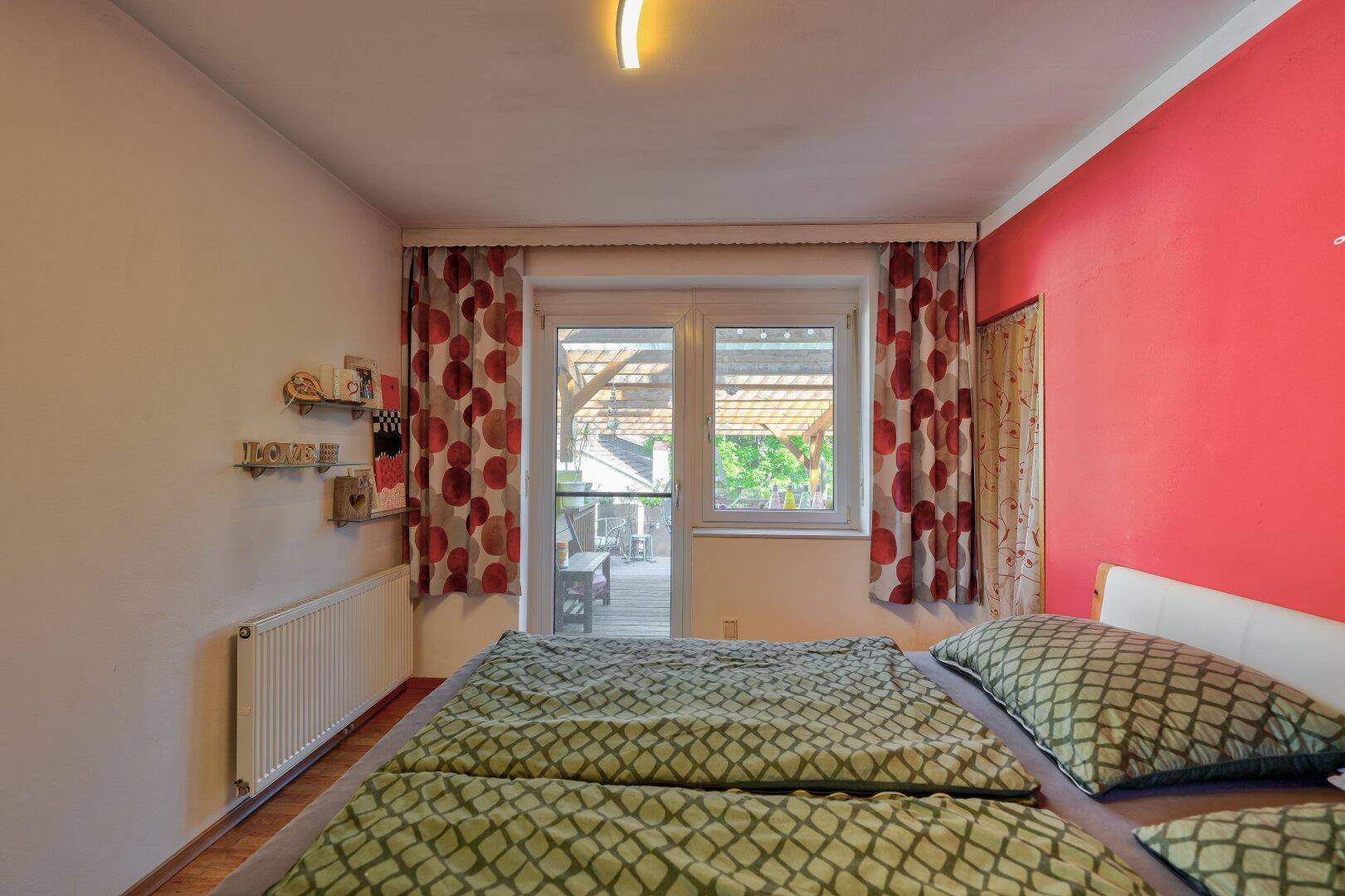 Schlafzimmer mit Doppelbett, roter Akzentwand und Zugang zur Terrasse.