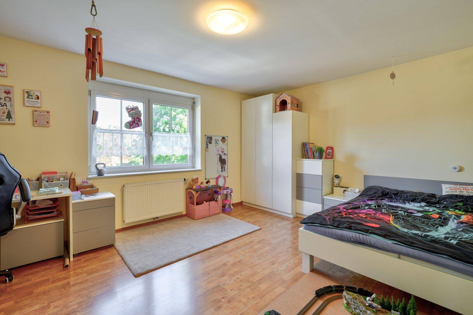 Helles Kinderzimmer mit Holzfußboden, Schreibtisch, Kleiderschrank und einem großen Fenster.