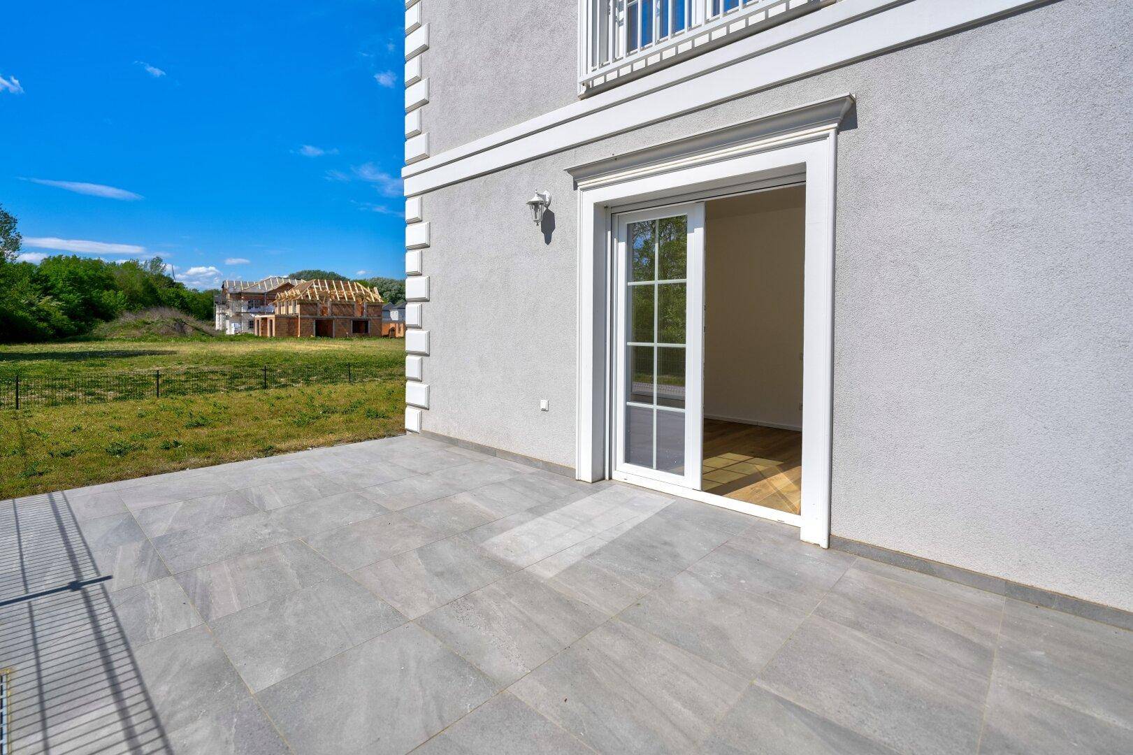Geräumige Terrasse mit grauen Fliesen und Zugang zum Innenbereich, umgeben von Grünflächen.