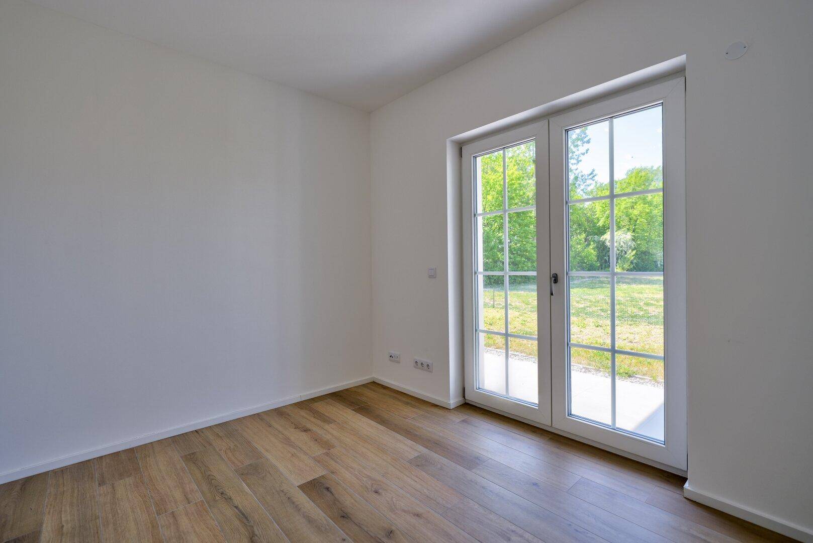 Leeres Zimmer mit Holzboden und großen Fenstertüren, die viel Licht hereinlassen.